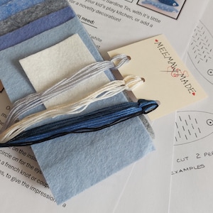 May include: A craft kit for a felt sardine tin, including stacked blue and grey felt squares, three skeins of embroidery floss in light grey, cream, and dark blue, and printed instruction sheets with the text MEEMAW MADE and Felt Sardine Tin.