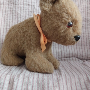 May include: A plush, light brown teddy bear with a peach-colored ribbon tied around its neck. The bear has small, dark eyes and a black nose. It is sitting on a pink and white striped fabric.