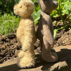 May include: Two handmade teddy bears stand side-by-side on a wooden surface. One is a light beige color with a fuzzy texture, and the other is brown with a tweed-like pattern. The bears are in a natural outdoor setting.