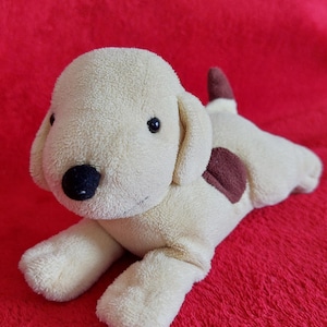 May include: A plush toy dog in a lying position with cream-coloured fur, a black nose, small black eyes, and brown patches on its back and tail. The stuffed animal features soft fabric and a simple design with floppy ears and a small stitched mouth.