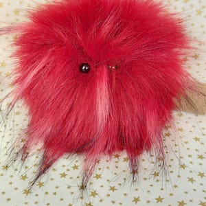 May include: A fluffy red and black plush toy with large black eyes. The toy is shaped like a ball with long, spiky fur.