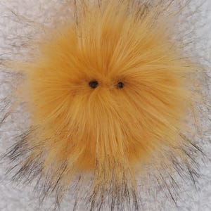 May include: A round, fluffy, yellow pompom with two dark brown button eyes. The pompom has long, soft, yellow fur with black tips, giving it a textured appearance. The background is a white, textured fabric.
