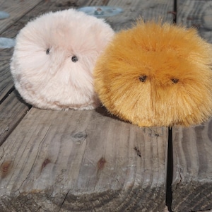May include: Two fluffy round plush toys with embroidered eyes. The toy on the left is light pink and the toy on the right is orange.