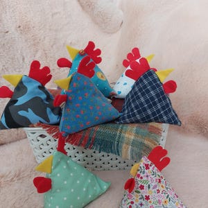 May include: A group of eight triangular fabric chicken cushions in various patterns. The cushions are made of different fabrics, including camouflage, polka dots, floral, and plaid. The cushions are all in a white basket.