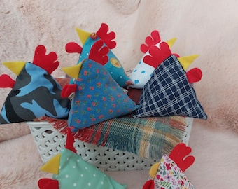 May include: A group of eight triangular fabric chicken pillows in various patterns. The pillows are made of different fabrics, including camouflage, polka dots, floral, and plaid. The pillows are all in a white basket.