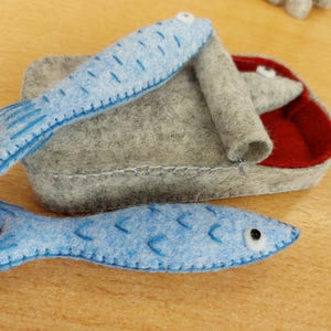 May include: Handmade felt toy set featuring two blue fish and a gray felt container with a red interior. The fish have stitched details and black eyes. The set is designed for children's play.