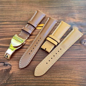 Può includere: Una collezione di cinturini per orologi in pelle in varie tonalità di marrone e beige, con cuciture bianche. Un cinturino presenta una chiusura color oro. I cinturini sono disposti su una superficie di legno scuro.