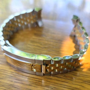 Può includere: Un cinturino per orologio bicolore caratterizzato da una combinazione di maglie in metallo color argento e color oro. Il cinturino include un meccanismo di chiusura pieghevole in metallo con un piccolo logo inciso sulla parte centrale.