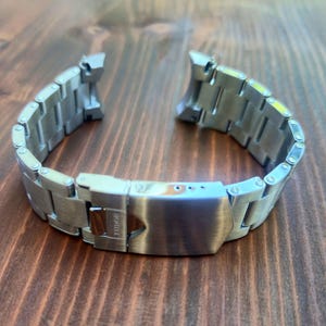 Peut inclure: Un bracelet de montre en acier inoxydable argenté avec une finition brossée. Le bracelet est ouvert, montrant le fermoir et les maillons. Le fermoir porte la marque "TUDOR". Il repose sur une surface en bois.