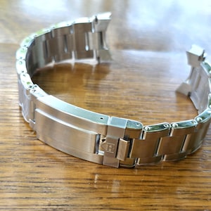 Può includere: Un cinturino per orologio in acciaio inossidabile color argento con finitura spazzolata, una chiusura deployante con un piccolo logo a scudo inciso e finali curvi progettati per adattarsi alla cassa dell'orologio.
