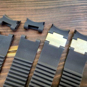 Puede incluir: Primer plano de varias correas y cierres de reloj negros. Las correas tienen un dise&ntilde;o acanalado y est&aacute;n hechas de un material flexible. Algunas correas tienen cierres de metal dorado. Los art&iacute;culos est&aacute;n dispuestos sobre una superficie de madera.