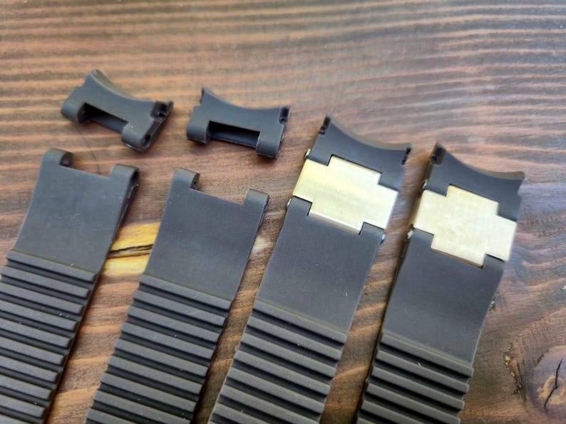 Puede incluir: Primer plano de varias correas y cierres de reloj negros. Las correas tienen un dise&ntilde;o acanalado y est&aacute;n hechas de un material flexible. Algunas correas tienen cierres de metal dorado. Los art&iacute;culos est&aacute;n dispuestos sobre una superficie de madera.