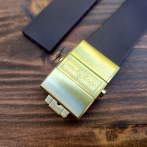 Może przedstawiać: Zbliżenie paska do zegarka ze złotym zapięciem. Zapięcie jest prostokątne z wygrawerowanym napisem "ULYSSE NARDIN". Pasek jest ciemnobrązowy i wygląda na sk&oacute;rzany.
