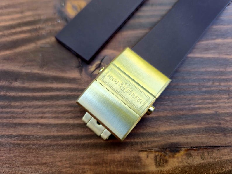 Może przedstawiać: Zbliżenie paska do zegarka ze złotym zapięciem. Zapięcie jest prostokątne z wygrawerowanym napisem "ULYSSE NARDIN". Pasek jest ciemnobrązowy i wygląda na sk&oacute;rzany.
