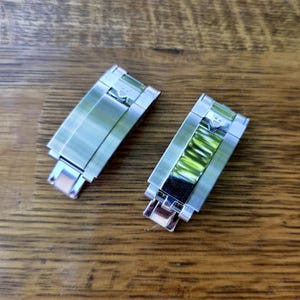 May include: Two silver-toned metal watch clasp links with a brushed finish. One link features a central decorative insert with a vibrant lime green, ribbed texture and a small shield-shaped emblem near the top hinge mechanism.