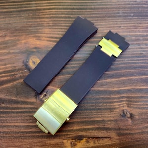 Puede incluir: Dos correas de reloj azul marino oscuro con cierres de metal color oro. Una correa está suelta, mientras que la otra está parcialmente ensamblada. Las correas están sobre una superficie de madera marrón oscuro.