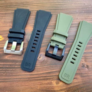 May include: Four watch straps in black and olive green are displayed on a wooden surface. Two straps are black, one with a silver buckle and the other with a black buckle. The other two straps are olive green, one with a black buckle.