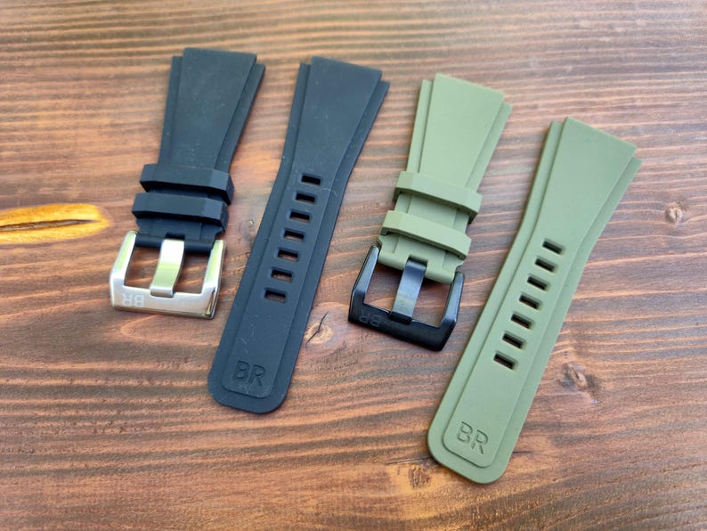 May include: Four watch straps in black and olive green are displayed on a wooden surface. Two straps are black, one with a silver buckle and the other with a black buckle. The other two straps are olive green, one with a black buckle.