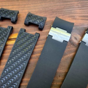 Puede incluir: Un conjunto de correas y cierres de reloj negros. Las correas tienen un patr&oacute;n de ondas texturizadas, y los cierres son plateados. Los art&iacute;culos est&aacute;n dispuestos sobre una superficie de madera.
