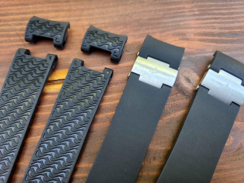 Puede incluir: Un conjunto de correas y cierres de reloj negros. Las correas tienen un patr&oacute;n de ondas texturizadas, y los cierres son plateados. Los art&iacute;culos est&aacute;n dispuestos sobre una superficie de madera.