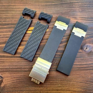 Puede incluir: Un conjunto de correas y cierres de reloj negros. Dos correas tienen un patr&oacute;n ondulado, mientras que las otras son lisas. Los cierres son dorados, dispuestos sobre una superficie de madera.