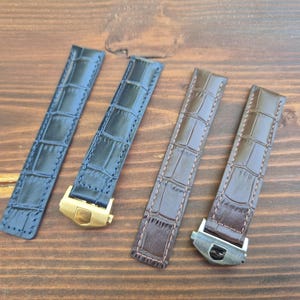 Könnte beinhalten: Vier Uhrenarmbänder in Blau- und Brauntönen mit Krokodillederstruktur. Die Armbänder haben Metallschnallen in Gold- und Silbertönen. Die Armbänder sind auf einer Holzoberfläche angeordnet.