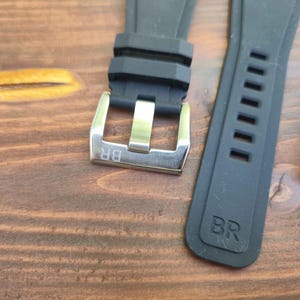 May include: Close-up of watch straps and buckles. The image shows a black rubber watch strap with a silver buckle, and a grey strap with rectangular cutouts. The buckles have the letters "BR" engraved.