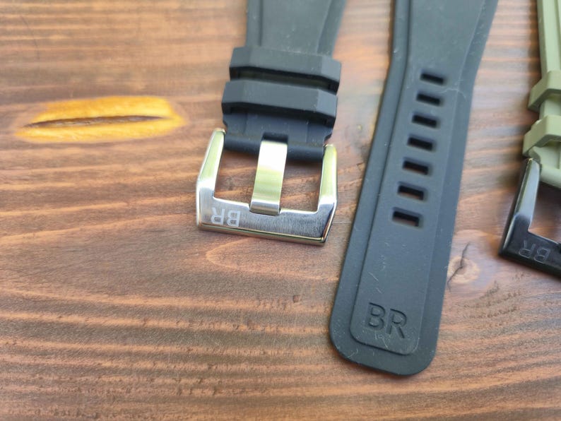 May include: Close-up of watch straps and buckles. The image shows a black rubber watch strap with a silver buckle, and a grey strap with rectangular cutouts. The buckles have the letters "BR" engraved.