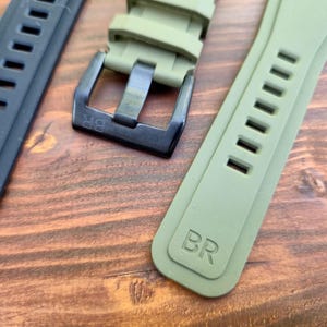 May include: Close-up of watch straps in black and olive green. The straps have a rectangular design with multiple holes and the letters "BR" embossed on them. The buckle is black.