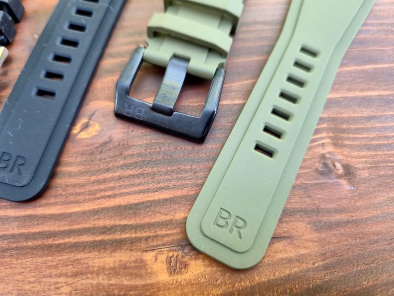 May include: Close-up of watch straps in black and olive green. The straps have a rectangular design with multiple holes and the letters "BR" embossed on them. The buckle is black.