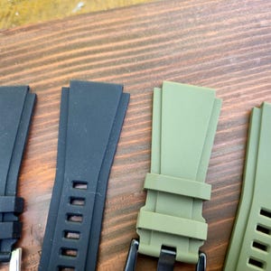 May include: Close-up of four watch straps in black and olive green. Two black straps have silver buckles and rectangular holes. The olive green straps have black buckles and rectangular holes. The straps are made of a flexible material.