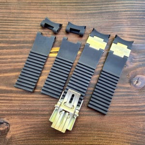 Puede incluir: Un conjunto de correas y conectores de reloj negros con cierres dorados, dispuestos sobre una superficie de madera. Las correas tienen un dise&ntilde;o acanalado y los conectores son peque&ntilde;os y rectangulares.
