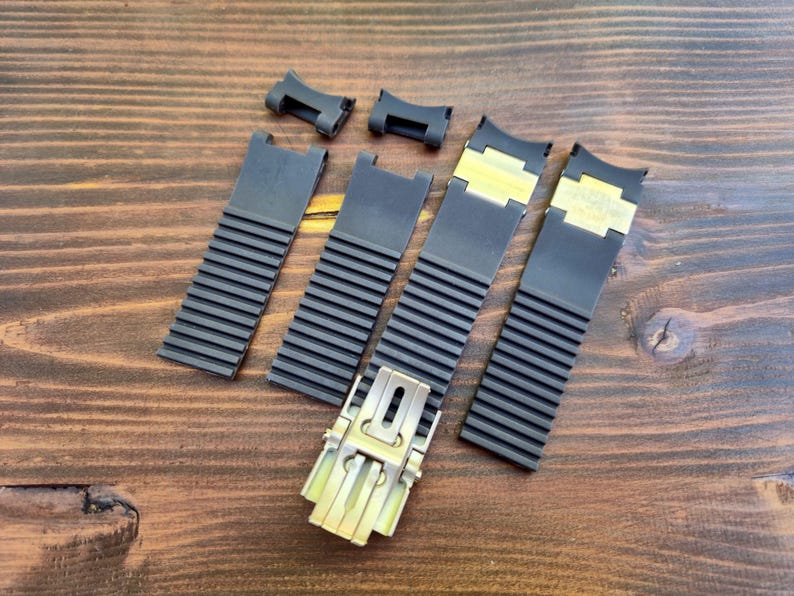 Puede incluir: Un conjunto de correas y conectores de reloj negros con cierres dorados, dispuestos sobre una superficie de madera. Las correas tienen un dise&ntilde;o acanalado y los conectores son peque&ntilde;os y rectangulares.