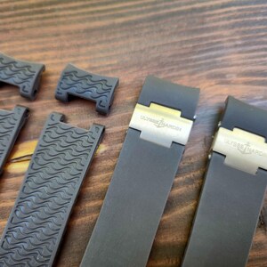 Puede incluir: Correas de reloj grises oscuras y piezas de conexi&oacute;n con un patr&oacute;n de ondas. Las correas tienen cierres plateados con el logotipo "ULYSSE NARDIN". Los art&iacute;culos se muestran sobre una superficie de madera.