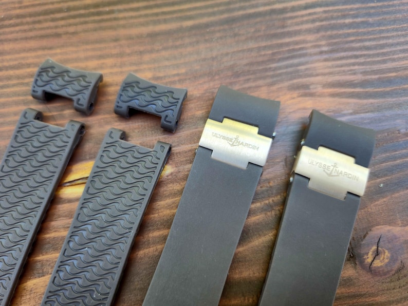 Puede incluir: Correas de reloj grises oscuras y piezas de conexi&oacute;n con un patr&oacute;n de ondas. Las correas tienen cierres plateados con el logotipo "ULYSSE NARDIN". Los art&iacute;culos se muestran sobre una superficie de madera.