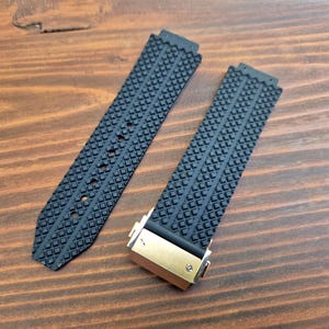 May include: Two black rubber watch straps with a textured, geometric pattern. One strap is attached to a gold-coloured clasp. The straps are displayed on a wooden surface.