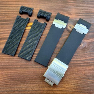 Puede incluir: Correas y componentes de reloj negros dispuestos sobre una superficie de madera. Las correas tienen un dise&ntilde;o texturizado y los cierres son plateados. Las correas son probablemente para un reloj de alta gama.