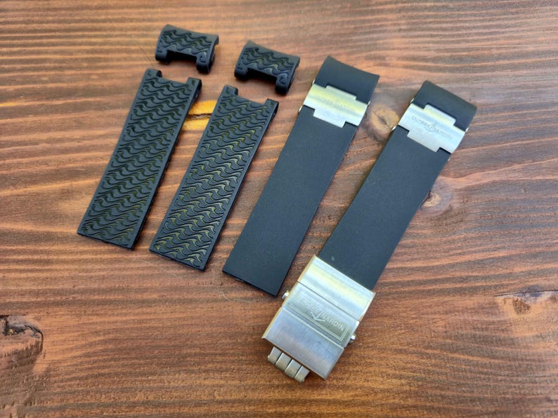 Puede incluir: Correas y componentes de reloj negros dispuestos sobre una superficie de madera. Las correas tienen un dise&ntilde;o texturizado y los cierres son plateados. Las correas son probablemente para un reloj de alta gama.