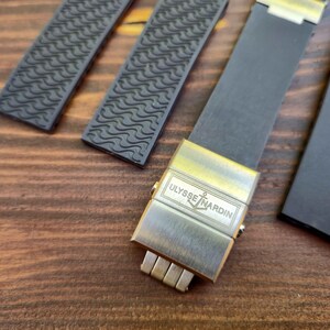Puede incluir: Primer plano de una correa de reloj con un cierre met&aacute;lico. La correa negra tiene un patr&oacute;n texturizado, y el cierre dorado presenta el logotipo "ULYSSE NARDIN". El fondo es una superficie de madera.