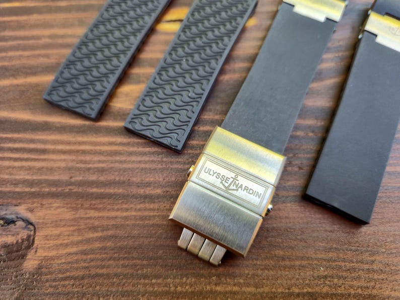 Puede incluir: Primer plano de una correa de reloj con un cierre met&aacute;lico. La correa negra tiene un patr&oacute;n texturizado, y el cierre dorado presenta el logotipo "ULYSSE NARDIN". El fondo es una superficie de madera.