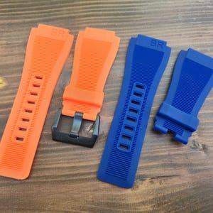 May include: Four watch straps in orange and blue. Two orange straps are next to a black buckle. The blue straps are on the right. All straps have the letters "BR" on them.