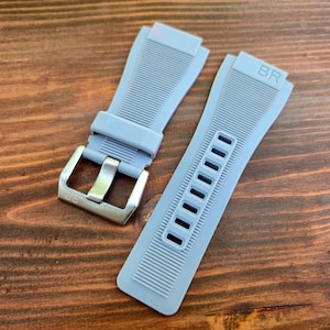 May include: Two light grey watch straps with a silver buckle and the letters "BR" embossed on one strap. The straps have a ribbed texture and rectangular cutouts. The watch straps are on a wooden surface.