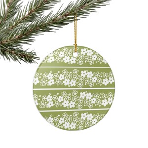 May include: A round, flat ceramic ornament featuring a sage green background with three horizontal bands of white, hand-drawn style floral patterns. The ornament is suspended by a thin gold cord and displays a repeating motif of small flowers.