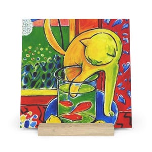 May include: A colorful art print featuring a yellow cat reaching into a fishbowl with red fish. The background includes a green garden scene, a red patterned wall, and a blue plate. The print is displayed on a wooden stand.