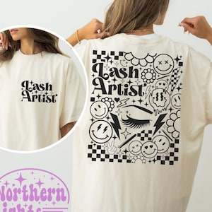 Puede incluir: Una camiseta blanca con un diseño gráfico negro en la espalda. El diseño presenta el texto "Lash Artist" rodeado de caras sonrientes, estrellas, flores y otros elementos decorativos. La parte delantera de la camiseta tiene el texto "Lash Artist" en negro.