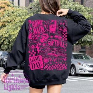 May include: Black sweatshirt featuring a bright pink graphic design. The design incorporates the phrases "Home Run", "Softball Vibes", and "Game Day", alongside baseball-related illustrations such as a baseball, a foam finger, and a bow. The sweatshirt has a crew neck and long sleeves.
