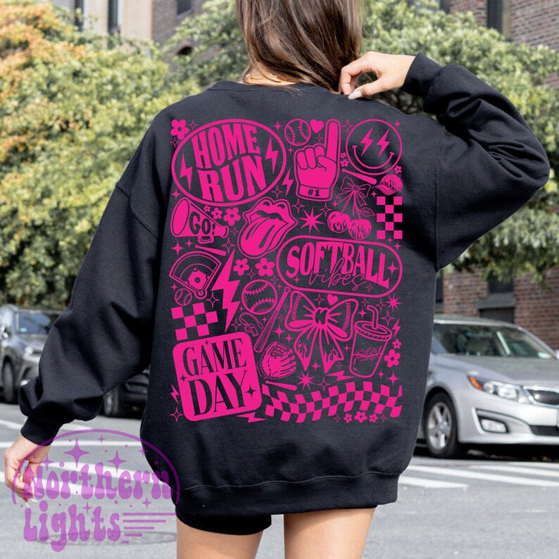 May include: Black sweatshirt featuring a bright pink graphic design. The design incorporates the phrases "Home Run", "Softball Vibes", and "Game Day", alongside baseball-related illustrations such as a baseball, a foam finger, and a bow. The sweatshirt has a crew neck and long sleeves.