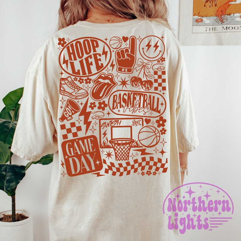 May include: Cream-colored t-shirt with a basketball-themed graphic in shades of brown. The design includes the words "HOOP LIFE," "BASKETBALL VIBES," and "GAME DAY," along with basketball-related imagery such as a basketball, hoop, and sneakers.