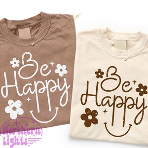 May include: Two t-shirts, one brown and one cream, with the words "Be Happy" in a whimsical font. The design includes flowers and a smiling face. Each shirt has a delicate gold necklace.
