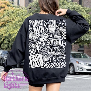 May include: Black sweatshirt with a white graphic design featuring softball-themed illustrations and text. The design includes the words "Home Run," "Softball Vibes," and "Game Day," along with baseballs, a bow, and a drink.